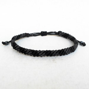 Puede incluir: Una pulsera trenzada negra con cierre de nudo corredizo ajustable. La pulsera está hecha de un material oscuro y tejido apretado, y presenta pequeñas cuentas oscuras en los extremos de los cordones. La pulsera se muestra sobre una superficie blanca.