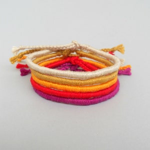 Könnte beinhalten: Fünf bunte Freundschaftsarmbänder in den Farben Rot, Orange, Gelb, Gold und Lila. Die Armbänder sind aus geflochtener Schnur gefertigt und miteinander verknotet.