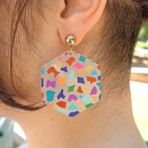 Puede incluir: Un par de pendientes hexagonales con un patrón de mosaico colorido. Los pendientes están hechos de acrílico transparente y tienen un poste dorado.