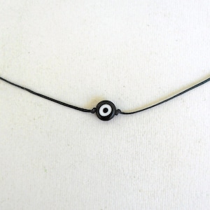 Può includere: Una collana in cordoncino di cuoio nero con un piccolo ciondolo di occhio turco nero con un centro bianco.
