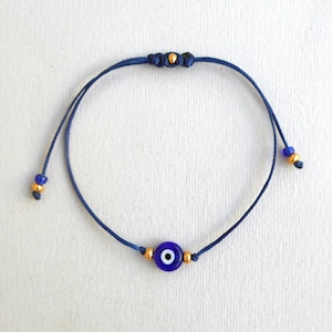 Op de afbeelding: Een donkerblauwe koordarmband met een centrale ronde glazen kraal in de vorm van een blauw, wit en zwart boze oog. De armband is voorzien van kleine goudkleurige tussenkralen aan weerszijden van de bedel en bij de uiteinden van het koord.