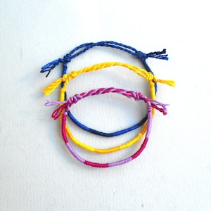 May include: Three friendship bracelets made of colorful string. The bracelets are blue, yellow, and pink.