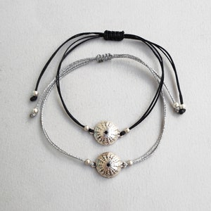 May include: Two bracelets with silver charms in the shape of a sea urchin. The top bracelet is black cord and the bottom bracelet is silver cord.