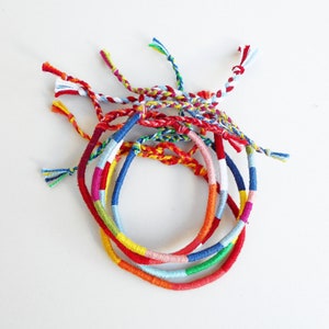 Könnte beinhalten: Ein Set bunter Freundschaftsarmbänder aus gewebtem Garn. Die Armbänder sind in Rot-, Blau-, Grün-, Gelb- und Weißtönen gehalten.