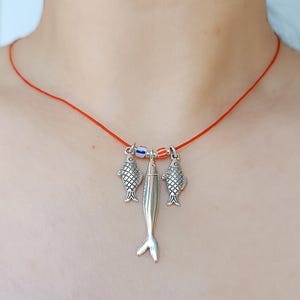 Op de afbeelding: Een ketting met een dun rood koord en drie zilverkleurige visbedels. De twee buitenste visbedels hebben een textuur met schubben, terwijl de middelste vis glad is. Tussen de bedels bevinden zich twee kleine gestreepte kralen.