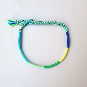 May include: A colorful friendship bracelet made with blue, green, and yellow thread. The bracelet has a tassel on one end.