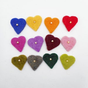 Puede incluir: Doce adornos de fieltro en forma de corazón en varios colores, incluyendo azul, amarillo, naranja y rojo. Cada corazón tiene una pequeña cuenta dorada en el centro. Los corazones están dispuestos sobre una superficie blanca.
