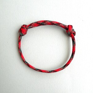 May include: A circular bracelet crafted from woven paracord featuring a vibrant camouflage pattern in shades of bright red, black, and grey. The cord is secured with two sliding knots on either side, allowing for adjustable sizing of the band.