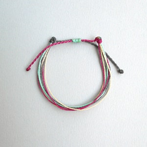 May include: A multi-colored friendship bracelet with grey, pink, yellow, and light blue threads. The bracelet is tied with a knot at each end.