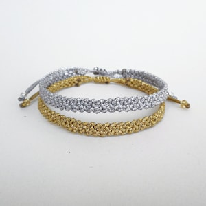 Può includere: Due braccialetti intrecciati, uno argentato e uno dorato, sono impilati l'uno sull'altro. I braccialetti sono realizzati con un cordoncino sottile e intrecciato.