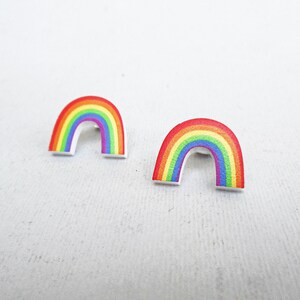 Puede incluir: Un par de pendientes de arcoíris en forma de arco. Los pendientes están hechos de madera y tienen un fondo blanco.