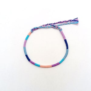 Könnte beinhalten: Ein farbenfrohes Freundschaftsarmband mit blauen, rosa und orangen Fäden. Das Armband ist mit einem Knoten gebunden und hat an einem Ende eine Quaste.