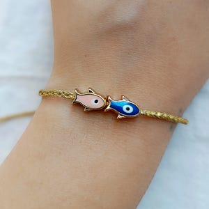 May include: A gold-toned braided cord bracelet featuring two small fish-shaped charms. One fish is light pink with a black eye, and the other is deep blue with a white and black eye, both designed with gold-toned metal outlines and details.