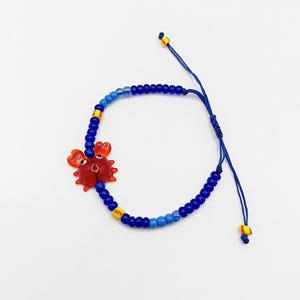 May include: A blue beaded bracelet with a red crab charm. The bracelet features a series of blue beads, with accents of yellow and light blue. The crab charm is red with black eyes and a smiling face.