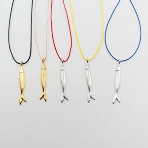 Puede incluir: Cinco collares con colgantes en forma de pez. Dos son dorados y tres plateados. Los collares tienen cordones de diferentes colores: negro, beige, rojo, amarillo y azul. Los colgantes tienen un diseño de cola de pez.