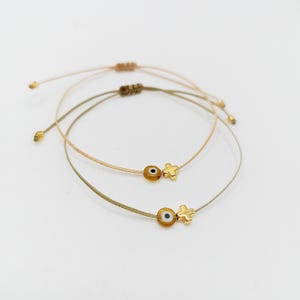 Könnte beinhalten: Drei verstellbare Armbänder mit Kordel. Zwei Armbänder haben eine goldfarbene Evil-Eye-Perle und einen goldfarbenen Kreuz-Anhänger. Das dritte Armband ist im Hintergrund sichtbar. Die Farben der Kordeln sind beige, olivgrün und hellbraun.