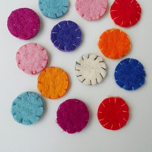 May include: A collection of colorful, felt circles in various shades of pink, blue, orange, red, purple, and white. Each circle is hand-stitched around the edge with contrasting thread, creating a decorative and textured appearance. The circles are arranged on a white surface.