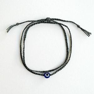 Könnte beinhalten: Ein schwarzes geflochtenes Armband mit einem blauen Glücksbringer in Form eines Auges.