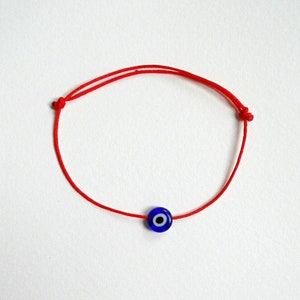 Può includere: Un braccialetto di corda rossa con un ciondolo blu a forma di occhio turco. Il ciondolo ha un centro bianco.