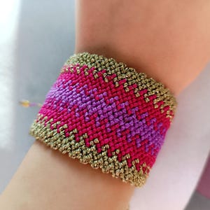 May include: A wide macrame bracelet in shades of pink and purple with a gold border. The bracelet features a geometric pattern and is worn on the wrist. The bracelet is handmade and has a bohemian style.