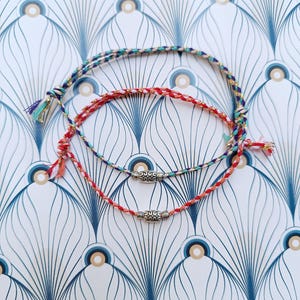 Könnte beinhalten: Drei farbenfrohe Freundschaftsarmbänder mit geflochtenen Kordeln in Rot, Türkis und Lila. Jedes Armband hat eine dekorative Silberperle und Quastenenden. Die Armbänder werden auf einem gemusterten Hintergrund präsentiert.