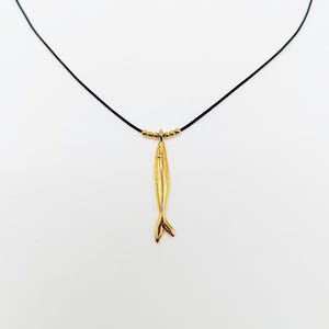 Puede incluir: Un collar con un colgante de pez dorado. El colgante en forma de pez cuelga de un cordón negro, con pequeñas cuentas doradas cerca de la parte superior. El colgante tiene un diseño detallado de aleta y cuerpo.