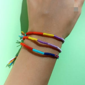 Puede incluir: Primer plano de una muñeca con tres pulseras de amistad de colores. Las pulseras están hechas de cuerda trenzada en tonos de rojo, amarillo, azul, morado y turquesa.