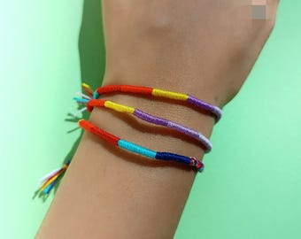 May include: A close-up of a wrist wearing three colorful friendship bracelets. The bracelets are made of woven string in shades of red, yellow, blue, purple, and turquoise.