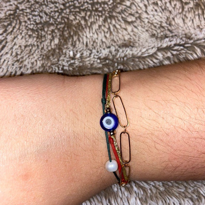 Op de afbeelding: Een set van gelaagde armbanden met een goudkleurige paperclipketting, een dun rood koord en een donkergroen koord. Het groene koord is voorzien van een blauw-witte boze oog kraal en een kleine witte, bolvormige kraal om de pols.