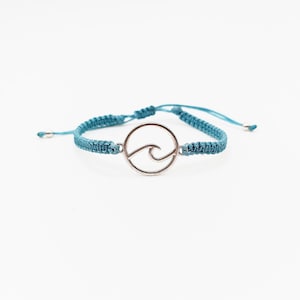 May include: A teal blue braided bracelet with a silver circle charm featuring a wave design.