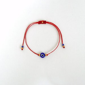 Op de afbeelding: Een rood koordarmbandje met een centrale blauwe, witte en zwarte boze oog kraal. Het armbandje bevat kleine goudkleurige metalen kralen aan weerszijden van de centrale bedel en aan de uiteinden van het verstelbare koord, dat ook blauwe kralen heeft.