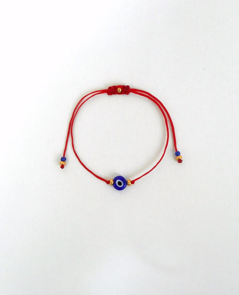 Op de afbeelding: Een rood koordarmbandje met een centrale blauwe, witte en zwarte boze oog kraal. Het armbandje bevat kleine goudkleurige metalen kralen aan weerszijden van de centrale bedel en aan de uiteinden van het verstelbare koord, dat ook blauwe kralen heeft.
