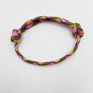 May include: A circular bracelet crafted from woven paracord featuring a camouflage pattern in vibrant shades of pink, olive green, and black. The cord is secured with two prominent knots on opposite sides, creating a simple and adjustable design.