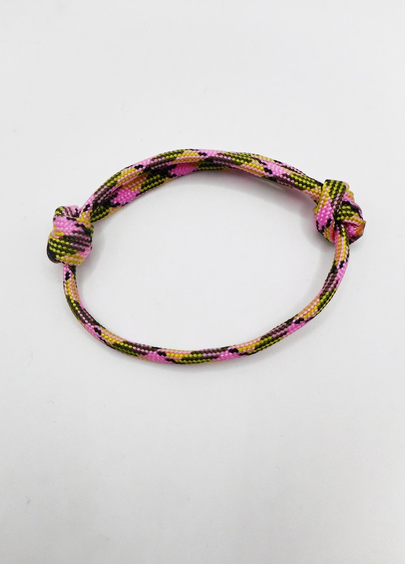 May include: A circular bracelet crafted from woven paracord featuring a camouflage pattern in vibrant shades of pink, olive green, and black. The cord is secured with two prominent knots on opposite sides, creating a simple and adjustable design.