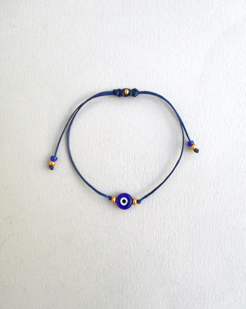 Op de afbeelding: Een donkerblauwe koordarmband met een centrale ronde glazen kraal in de vorm van een blauw, wit en zwart boze oog. De armband is voorzien van kleine goudkleurige tussenkralen aan weerszijden van de bedel en bij de uiteinden van het koord.