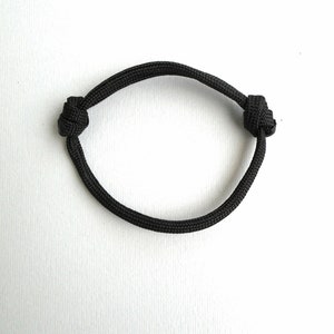 May include: A black cord bracelet constructed from a single piece of woven nylon material. The bracelet features two adjustable sliding knots on opposite sides, allowing the circular band to expand or contract for a secure fit around the wrist.