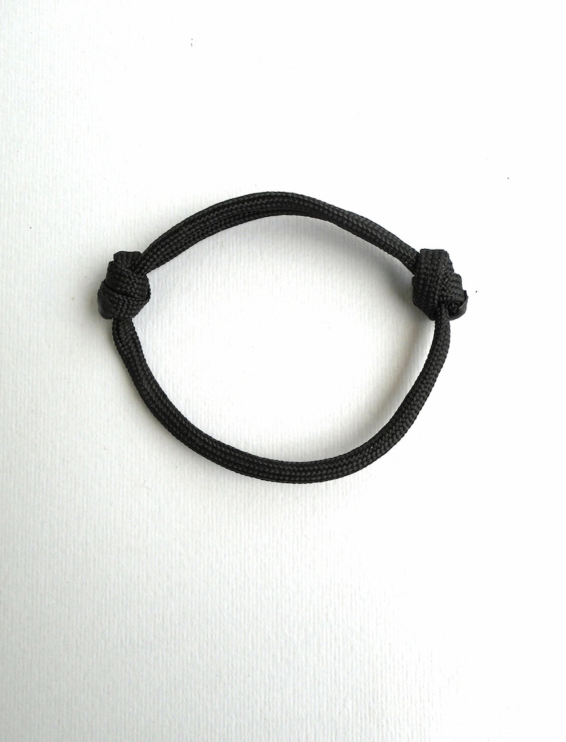 May include: A black cord bracelet constructed from a single piece of woven nylon material. The bracelet features two adjustable sliding knots on opposite sides, allowing the circular band to expand or contract for a secure fit around the wrist.