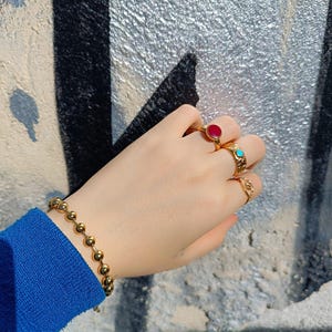 May include: A gold-toned ball chain bracelet and three gold-toned rings. One ring features a round red stone, another has a small turquoise inlay, and the third is a simple band with a cutout design. The jewelry is worn on a wrist with a blue sleeve.