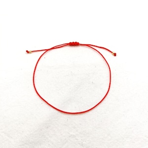 May include: A thin red cord bracelet featuring an adjustable sliding knot mechanism in the centre and two small gold-toned metal beads at the ends of the pull strings. The bracelet is constructed from a single continuous strand of red thread.
