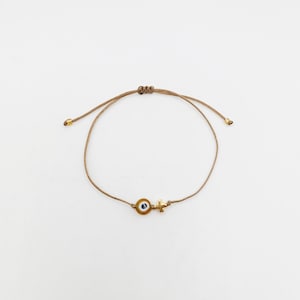 May include: A tan cord bracelet with a gold-coloured cross charm and an evil eye charm. The bracelet has adjustable sliding knots and gold-coloured end beads. The bracelet is displayed on a white background.