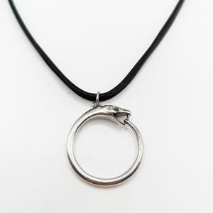 May include: A silver-toned metal pendant shaped like a snake biting its own tail to form a circular loop, suspended from a smooth black cord. The snake head is detailed with visible eyes and fangs, creating a continuous Ouroboros-style design.