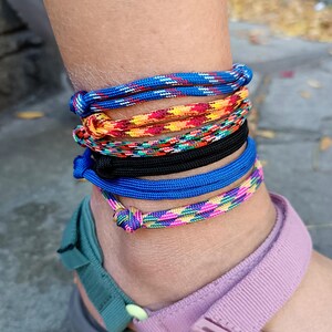 May include: A set of five stacked ankle bracelets made from woven cord. The bracelets feature various patterns and colors, including blue with white flecks, a multi-colored warm-toned weave, a black cord, a solid blue cord, and a pink and green pattern.