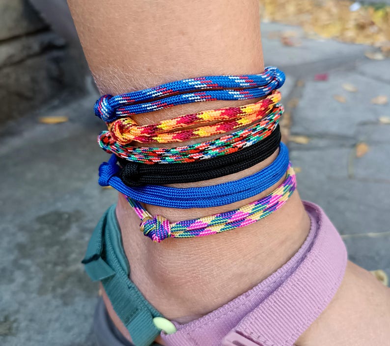 May include: A set of five stacked ankle bracelets made from woven cord. The bracelets feature various patterns and colors, including blue with white flecks, a multi-colored warm-toned weave, a black cord, a solid blue cord, and a pink and green pattern.