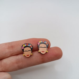 Puede incluir: Un par de pendientes de madera con un retrato de Frida Kahlo con una corona de flores. Los pendientes tienen detalles dorados.