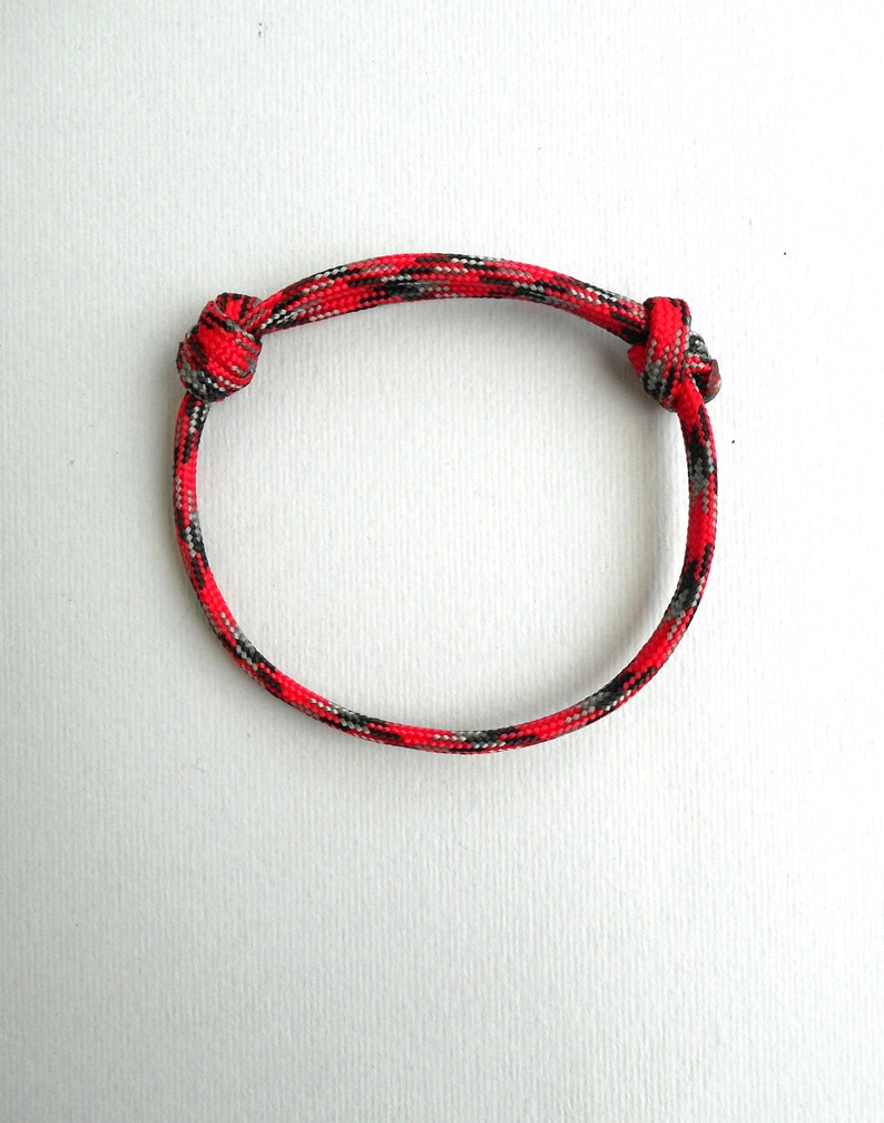 May include: A circular bracelet crafted from woven paracord featuring a vibrant camouflage pattern in shades of bright red, black, and grey. The cord is secured with two sliding knots on either side, allowing for adjustable sizing of the band.