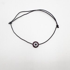 Puede incluir: Un brazalete de cuerda negra con un amuleto de ojo turco azul oscuro. El amuleto tiene un centro blanco y una pupila negra.