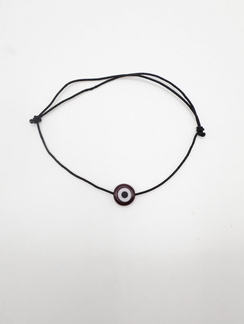 Puede incluir: Un brazalete de cuerda negra con un amuleto de ojo turco azul oscuro. El amuleto tiene un centro blanco y una pupila negra.