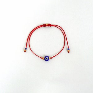 Op de afbeelding: Een rood koordarmbandje met een centrale blauwe, witte en zwarte kraal in de vorm van een boze oog. Het armbandje bevat kleine goudkleurige metalen kralen naast het centrale oog, met extra blauwe en gouden kralen aan de uiteinden van de koordjes.