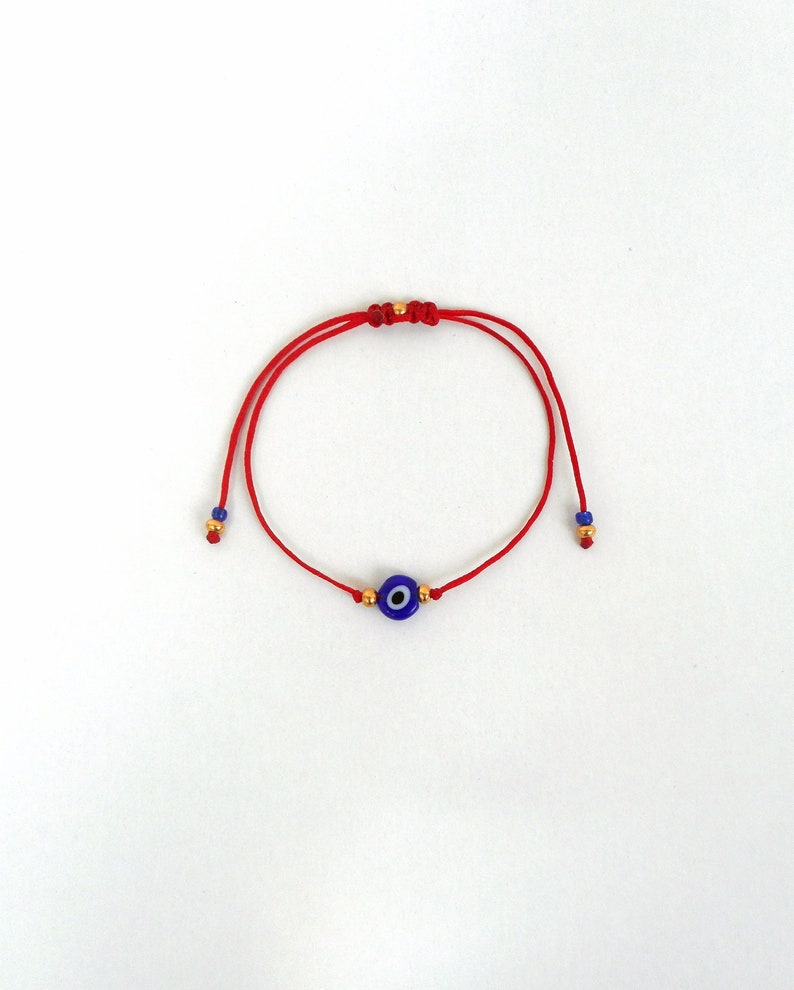 Op de afbeelding: Een rood koordarmbandje met een centrale blauwe, witte en zwarte kraal in de vorm van een boze oog. Het armbandje bevat kleine goudkleurige metalen kralen naast het centrale oog, met extra blauwe en gouden kralen aan de uiteinden van de koordjes.