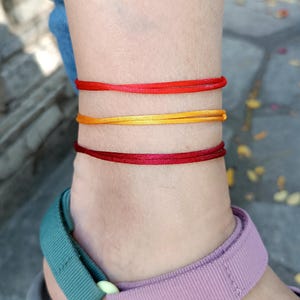 May include: Three thin cord anklets worn around the ankle. The top anklet is bright red, the middle anklet is golden yellow, and the bottom anklet is a deep burgundy red. Each anklet consists of a smooth, slender string material wrapped around.